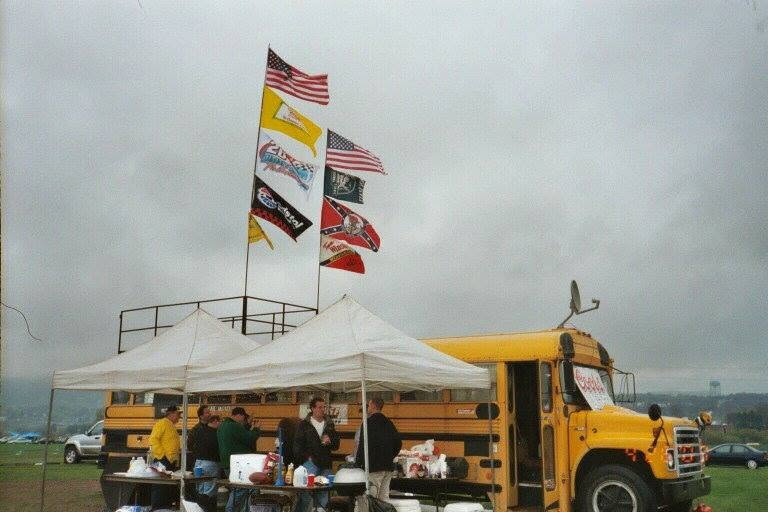 The big yellow tailgate bus with a roof deck, a tall pole full of flags, a satellite dish, two white pop-up tents, and the crew hanging out on an overcast day.